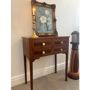 May include: A wooden console table with four drawers featuring round brass knobs. On top sits a brass candlestick with a lit candle and a framed floral painting in a dark ornate frame. Beside the table is a tall wooden pedestal with a potted plant.