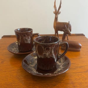 May include: Two ceramic espresso cups and matching saucers featuring a dark brown glaze with a white drip pattern near the rims. The cups have a cylindrical shape with a single handle, and the saucers have a matching brown and white glaze.