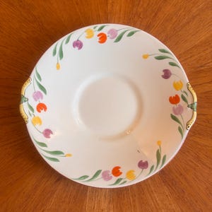 Może przedstawiać: Biały ceramiczny talerz do serwowania z kwiatowym wzorem tulipanów w kolorze pomarańczowym, żółtym i fioletowym. Talerz ma centralne zagłębienie i złote uchwyty. Zielone liście zdobią kwiaty.