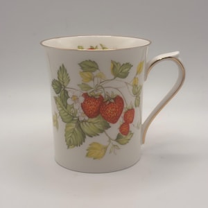 May include: White ceramic mug with a gold rim and handle, decorated with a strawberry plant design. The design features red strawberries, green leaves, and white flowers. The mug has a curved handle.