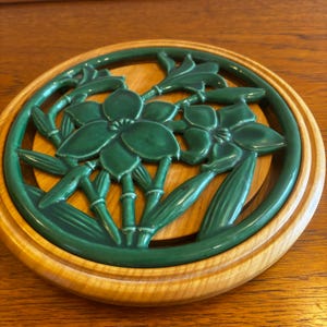 Può includere: Un sottopentola rotondo con un inserto in ceramica smaltata verde scuro caratterizzato da un design in rilievo di due fiori e steli simili a bambù, inserito in una cornice di legno chiaro e liscia con venature visibili e bordo circolare.