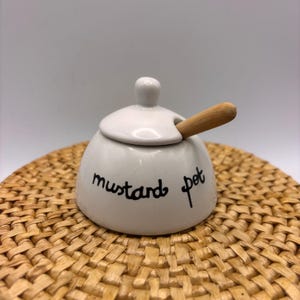 Può includere: Un vasetto di senape in ceramica bianca con coperchio e un piccolo cucchiaio di legno. Le parole "mustard pot" sono scritte in nero sul davanti. Il vasetto è appoggiato su un sottobicchiere intrecciato.