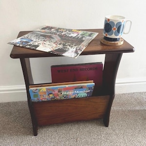 May include: A dark wood side table with a magazine rack base, holding a vinyl record sleeve on top and two records inside. A white ceramic mug with a blue and orange floral pattern sits on a wooden coaster on the table surface.