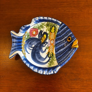 May include: A ceramic fish-shaped dish with a hand-painted design. The dish features a blue and white colour scheme with a detailed interior design of flowers and abstract shapes. The fish has a yellow beak and a blue striped body.