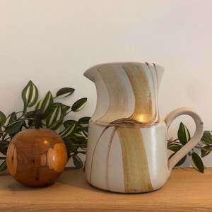 Può includere: Brocca in ceramica con base color crema e strisce verticali beige. A sinistra della brocca c'è un oggetto decorativo in legno a forma di mela. Piante verdi e frondose sullo sfondo. Oggetto decorativo.