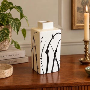 May include: A square ceramic vase with a white base and black paint splatter patterns. The vase features a narrow neck and a flat top opening. The design consists of bold, irregular black lines and dots applied across the white surface of the vessel.