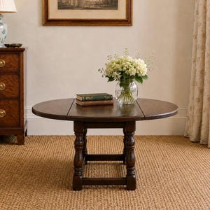 May include: A round, dark brown wooden drop-leaf table with a smooth surface and turned legs connected by a rectangular base. Two stacked books and a clear glass vase filled with white hydrangea flowers sit on the centre of the tabletop.