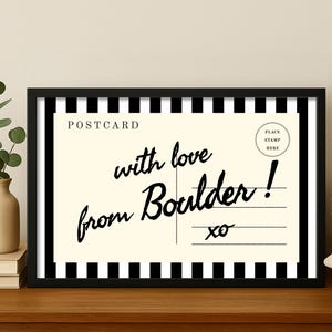 May include: A framed postcard print with a black and white striped border. The postcard has the words "with love from Boulder!" in a handwritten font. The print is displayed on a wooden surface.