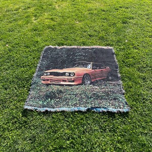 May include: Woven throw blanket featuring a vintage orange car design. The car is depicted in a forest setting. The blanket has a fringed edge and is displayed on a green grassy surface.