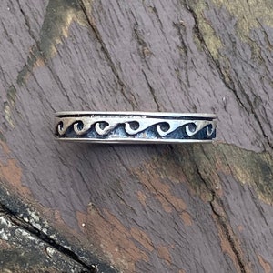 May include: A silver band ring with a wave design. The ring is on a wooden surface.