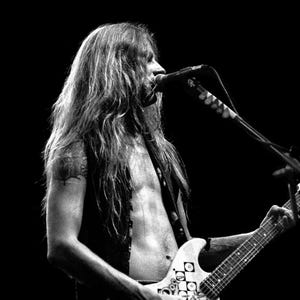 May include: Black and white photo of a musician performing on stage. The musician is playing an electric guitar with a unique design. The guitar has a white body with black and white geometric patterns. The musician is singing into a microphone.