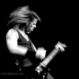 Puede incluir: Imagen en blanco y negro de una persona tocando una guitarra el&eacute;ctrica. La persona lleva una camiseta sin mangas negra y tiene el pelo largo. La imagen est&aacute; borrosa, lo que sugiere movimiento. El fondo es negro.