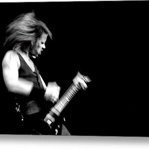 Puede incluir: Imagen en blanco y negro de una persona tocando una guitarra el&eacute;ctrica. La persona lleva una camiseta sin mangas negra y tiene el pelo largo. La guitarra est&aacute; enfocada, con la cara y el pelo borrosos por el movimiento.