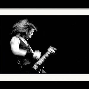 Puede incluir: Fotograf&iacute;a enmarcada en blanco y negro de una persona tocando una guitarra el&eacute;ctrica. La persona lleva una camiseta sin mangas y tiene el pelo largo. La imagen es un primer plano, con la persona y la guitarra enfocadas sobre un fondo oscuro.