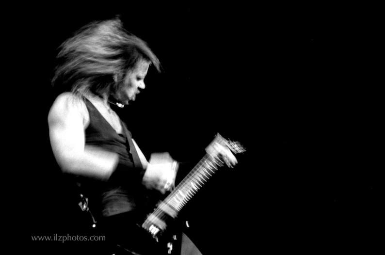 Puede incluir: Imagen en blanco y negro de una persona tocando una guitarra el&eacute;ctrica. La persona lleva una camiseta sin mangas negra y tiene el pelo largo. La imagen est&aacute; borrosa, lo que sugiere movimiento. El fondo es negro.