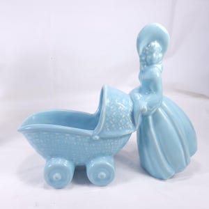Puede incluir: Un conjunto de figuras de cerámica azul claro con una mujer con un vestido largo y sombrero, junto a un cochecito de bebé. El cochecito tiene cuatro ruedas y un diseño de cesta tejida. El conjunto es un artículo decorativo vintage.