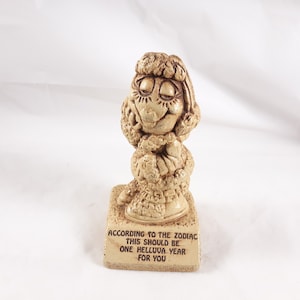 May include: A ceramic figurine of a woman with a large, curly hairstyle. The figurine is standing on a base with the text "According to the Zodiac this should be one helluva year for you".