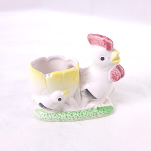 May include: A ceramic egg cup in the shape of a white rooster with a red comb and yellow beak. The egg cup is a yellow eggshell. The base is green with a small chick. A charming kitchen decor item.