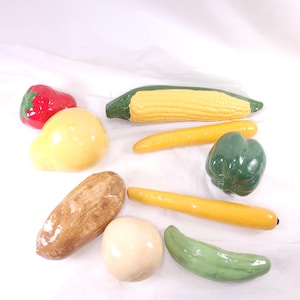 May include: A collection of ceramic vegetables, including a red pepper, corn on the cob, carrots, a green bell pepper, a cucumber, a potato, a pear, and a white onion. The vegetables are painted with realistic colors and details.