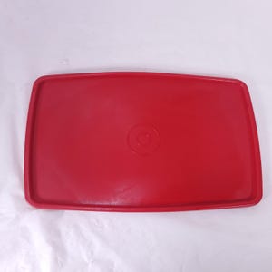 May include: A rectangular, red plastic lid with rounded corners. The lid has a slightly raised center with a circular indentation. The overall design is simple and functional, suitable for food storage containers.