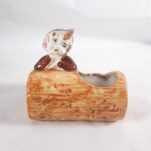 May include: A ceramic planter shaped like a log with a small figurine of a person peeking out from the top. The planter is brown and has a textured surface.