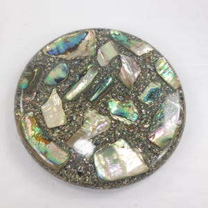 May include: A circular coaster featuring a mosaic of iridescent abalone shell fragments embedded in a dark, speckled resin base. The shell pieces display varied colors including green, blue, pink, and white, creating a reflective surface.