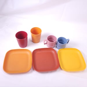 May include: A collection of colorful tableware, including plates, cups, and mugs. The set features square plates in orange, red-orange, and yellow. There are also red and orange cups, and pink and blue mugs with handles. The items are arranged on a white surface.
