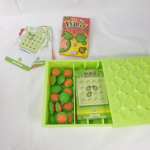 May include: A Flip It board game set featuring a bright lime green plastic grid tray, a stack of challenge cards, an instruction booklet with a turtle illustration, and a collection of small, round, orange and green turtle-shaped game pieces.