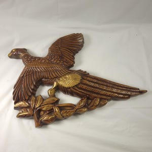 May include: A wooden wall hanging in the shape of a pheasant with outstretched wings. The bird is primarily brown with gold accents on the wings and a laurel wreath. The pheasant is a decorative item.