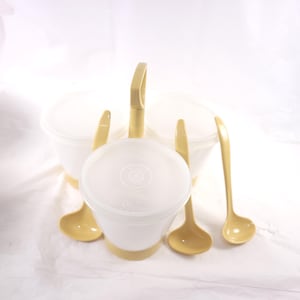 May include: A vintage kitchen set featuring three white containers with lids, a yellow handle, and three yellow ladles. The containers have a yellow base. The set is designed for serving or storing food.