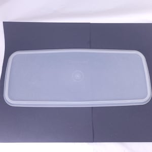 May include: A clear, rectangular Tupperware lid. The lid is made of a translucent, light-blue plastic and has a slightly raised edge. The Tupperware logo is visible on the lid. The lid is designed to fit a rectangular container.