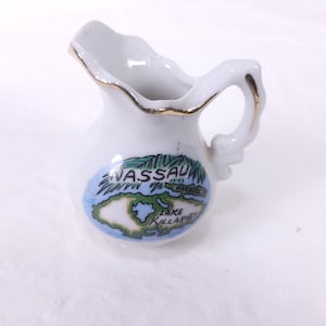 May include: Small white ceramic creamer with a gold rim and handle. The creamer features a colorful illustration of Nassau, Paradise Island, and Lake Killarney. A decorative souvenir item.
