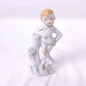 May include: A porcelain figurine of a young boy in a white outfit with gold trim, leaning against a white tree trunk. The boy has blonde hair and is posed with one hand on his hip. The figurine has gold accents on the tree trunk and base.