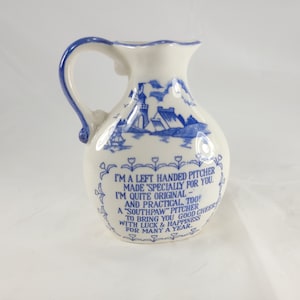 May include: A white ceramic pitcher with a blue handle and rim. The pitcher features a blue landscape design and text that reads "I'M A LEFT HANDED PITCHER MADE 'SPECIALLY FOR YOU." A decorative and functional kitchenware item.