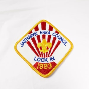 May include: A white diamond-shaped patch with a yellow border. The patch has red and white stripes with a yellow fleur-de-lis in the center. The text "Jayhawk Area Council" is in blue and the text "Lock In 1993" is in red.