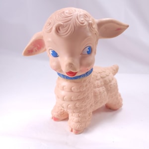 May include: A vintage, light pink lamb toy with blue eyes, a red mouth, and a blue collar. The lamb has curly hair and a textured body. The toy is standing on four legs and has pink accents on its ears and feet.
