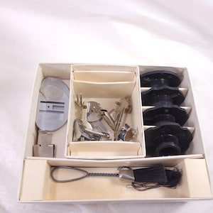 May include: A vintage sewing machine attachment set in a cream-colored box. The set includes various metal presser feet, black bobbins, and a silver-toned tassel. The box is divided into compartments for organized storage of the sewing accessories.