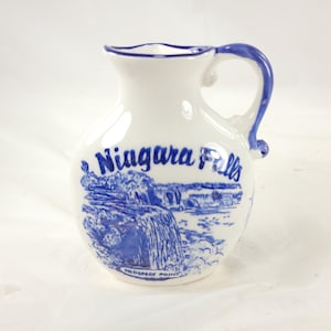 May include: White ceramic pitcher with a blue handle and rim. The pitcher features a blue illustration of Niagara Falls with the words "Niagara Falls" and "Prospect Point" in blue lettering.