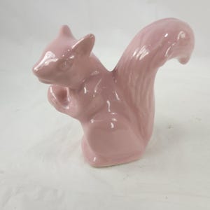 May include: A pink ceramic squirrel figurine. The squirrel is in a seated position, holding something in its paws, with a curved tail. The figurine has a glossy finish and is a solid, soft pink color.