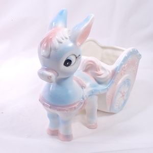 May include: A ceramic donkey planter in pastel blue, pink, and white. The donkey is pulling a decorative cart with floral accents. The donkey has large eyes and a sweet expression, perfect for a nursery or a whimsical touch to any room.