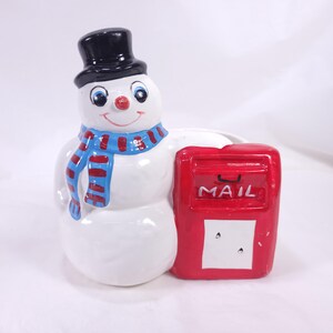 May include: A ceramic snowman figurine with a black top hat, blue and red striped scarf, and a red nose. The snowman is next to a red mailbox with the word "MAIL" on it. The figurine is white with blue and red accents.