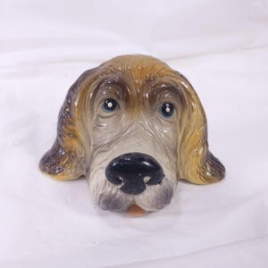 May include: A ceramic dog head sculpture with a detailed, hand-painted design. The dog has brown and tan fur, blue eyes, and a black nose. The sculpture is a decorative item.