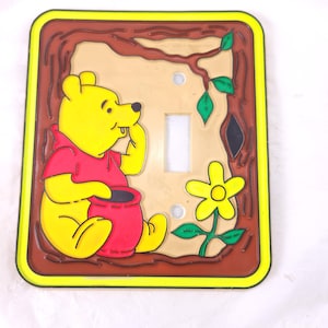 May include: Yellow and red Winnie the Pooh light switch cover. The cover features a yellow border, a brown tree design, and a light switch opening. Winnie the Pooh is wearing a red shirt and holding a red pot. A yellow flower and green leaves are also present.
