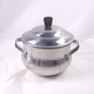 May include: A stainless steel lidded pot with two handles and a decorative finial. The pot has a rounded body and a lid with a small, ornate handle. The handles are decorated with a dark, patterned design.
