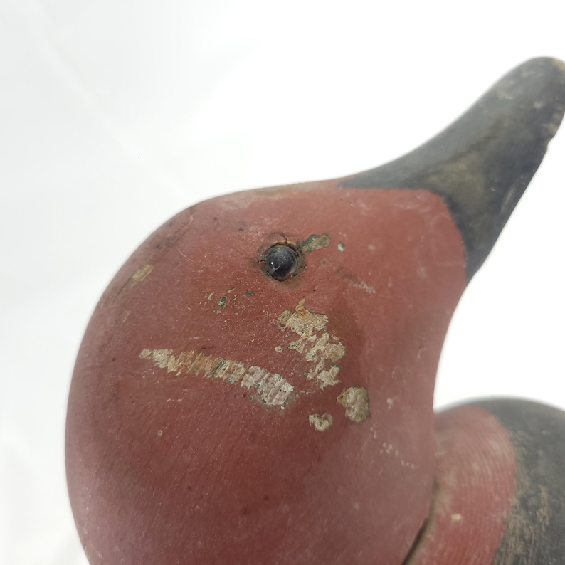 May include: Close-up of a vintage wooden duck decoy. The duck has a red head and neck with a black beak. The paint shows signs of wear, adding to its antique charm. A classic piece of folk art.