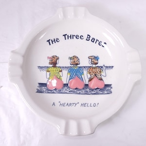 May include: White ceramic ashtray with a scalloped edge. The ashtray features a cartoon illustration of three figures with peach-shaped bottoms and the text "The Three Bares" and "A "Hearty" Hello!"