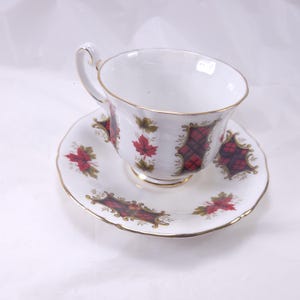 May include: White teacup and saucer set with gold trim. The cup and saucer feature a red and black plaid pattern with red maple leaves and gold accents. The set is designed for tea.