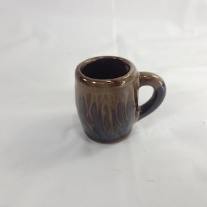 May include: A small ceramic mug with a brown and dark blue glaze. The mug has a rounded body, a dark interior, and a handle. The glaze has a dripped effect, creating a unique texture. The mug is suitable for espresso or small beverages.