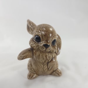 May include: A ceramic bunny figurine in shades of brown. The bunny has large, dark eyes, a small black nose, and detailed fur texture. Its ears are perked up, and it has one arm raised, giving it a playful appearance. A decorative item.
