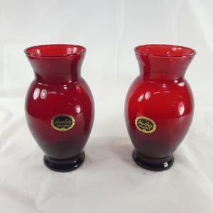 May include: Two matching ruby red glass vases with a bulbous body and a flared rim. Each vase features a circular gold and black sticker on the front that reads Royal Ruby Anchorglass, indicating the brand and origin of these glass vessels.